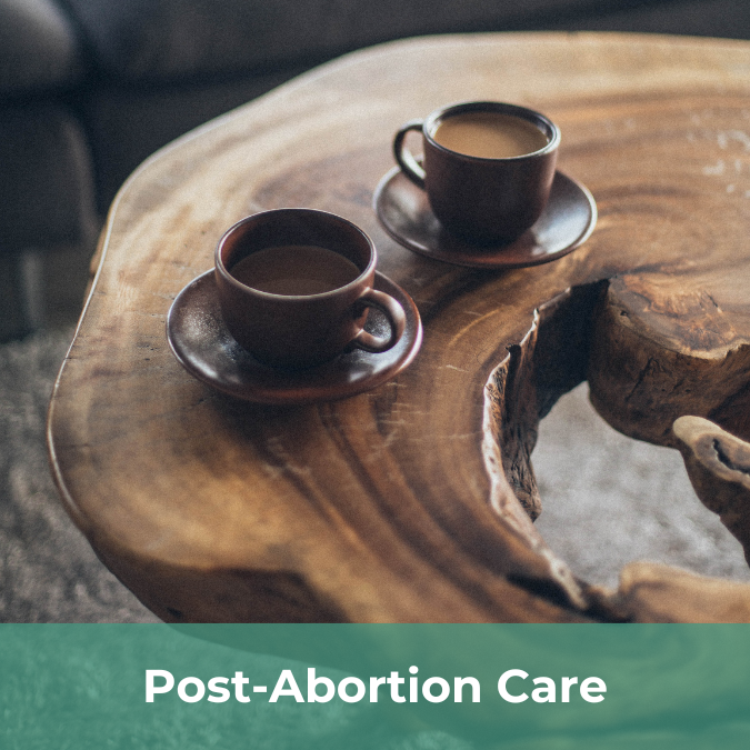 Two cups of coffee rest on a live edge would table. There is a text bar on the bottom of the image that reads Post-Abortion Care.